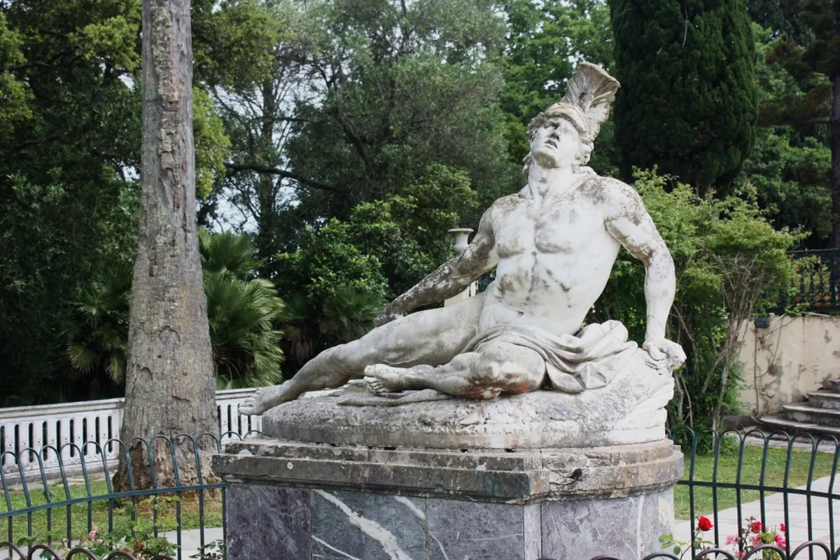 Una statua in marmo bianco di un guerriero ferito con elmo crestato, reclinata su una base di pietra tra gli alberi del giardino (l'Achille morente al palazzo Achilleion a Corfù, non lo spettacolo)
