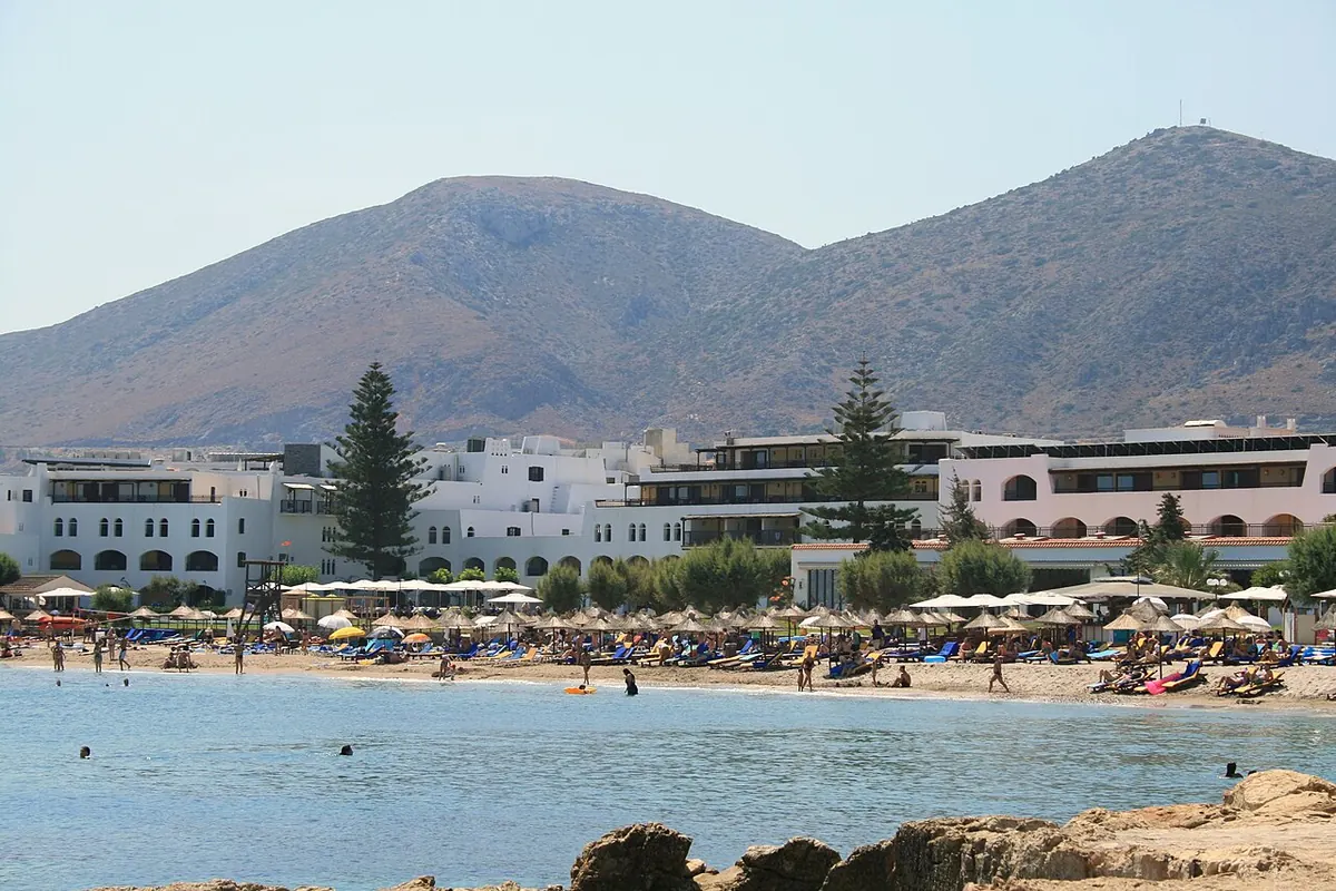 Una spiaggia sabbiosa con ombrelloni sotto bianchi edifici alberghieri e una montagna bruna alle spalle (la spiaggia di Hersonissos sulla costa nord di Creta, non il teatro)