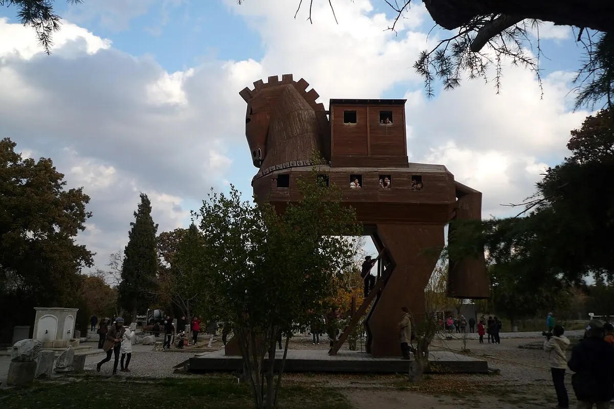 Un alto cavallo di legno con una piattaforma simile a una fortezza sul dorso si erge tra i pini sotto un cielo nuvoloso, con visitatori che camminano ai suoi piedi (la replica del cavallo di Troia nel sito archeologico di Troia in Turchia, non parte dello spettacolo)